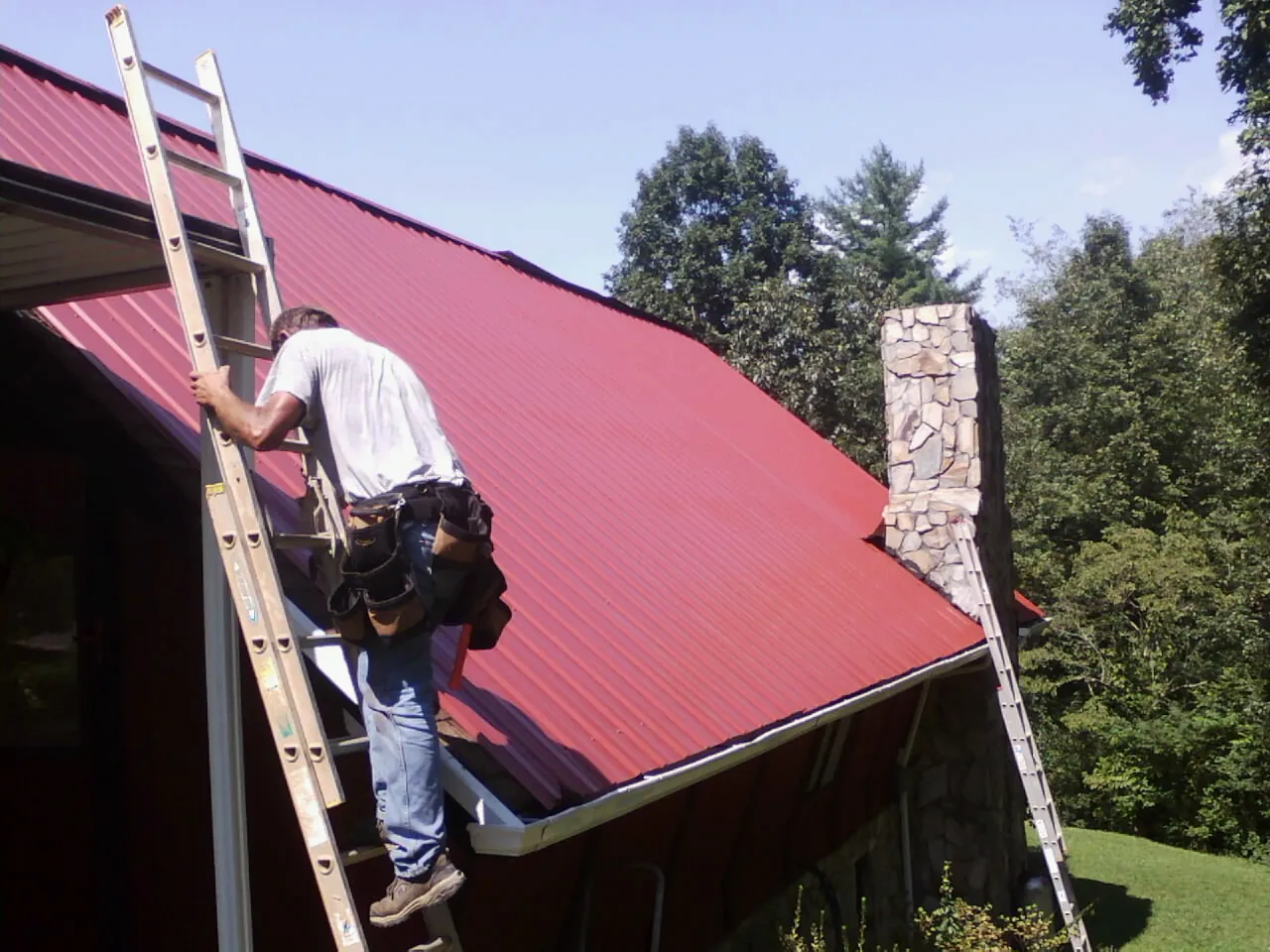 Roofer on a ladder beside a red metal roof and stone chimney — Soffit & Fascia Repair by Roofing Brothers in West Alexandria